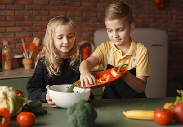 Little girl and boy mixing cutted pepper with other fresh ingredients for salad in big bowl while cooking at table with various fresh vegetables and fruits