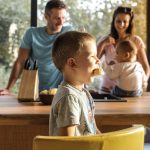 Side view of playful boy eating cookie while sitting at table with parents and sibling on background spending time at home