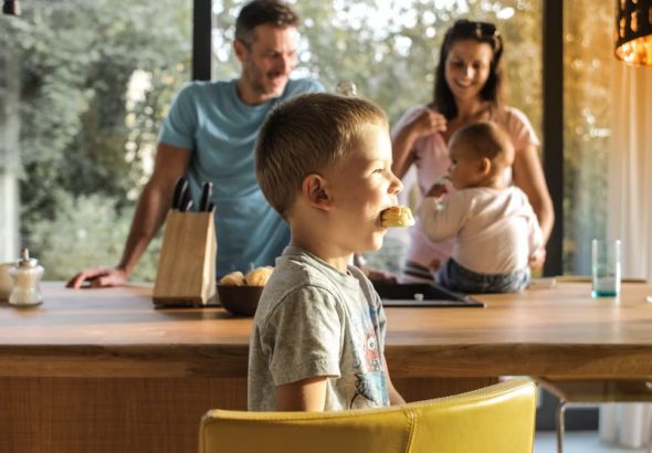 Side view of playful boy eating cookie while sitting at table with parents and sibling on background spending time at home